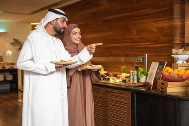 Delicious and fresh foods and pastries, a variety of different dishes, choosing a breakfast dish, enjoying breakfast, spending enjoyable times on the weekend, the concept of a happy family, an Emirati Arab couple wearing traditional Emirati attire serving food from the open buffet. Delicious and fresh foods and pastries, a variety of different dishes, choosing a breakfast dish, enjoying breakfast, spending enjoyable times on the weekend, the concept of a happy family, an Emirati Arab couple wearing traditional Emirati attire serving food from the open buffet.