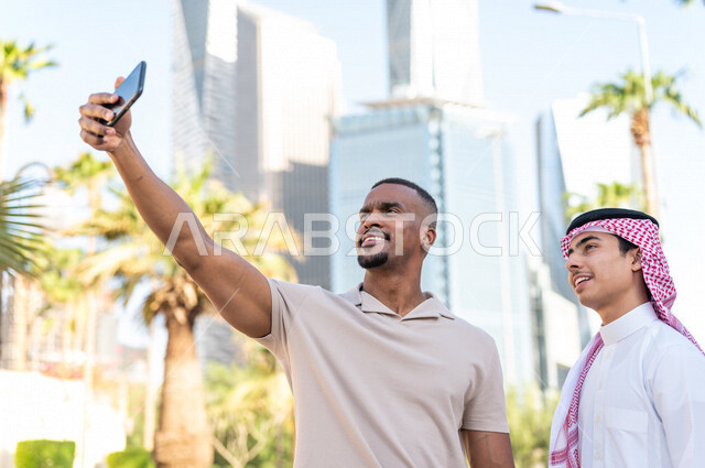 شابان عربيان خليجيان سعوديان مبتسمان يقفان بجانب بعضهما و يقومان بإلتقاط صورة سيلفي ، صورة تذكارية ، إجتماع صداقة ، قضاء وقت ممتع في مدينة الملك عبد الله المالية ( كافد ) في الرياض في المملكة العربية السعودية