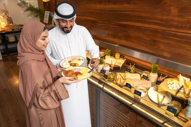 اطباق متنوعه ومختلفه من المأكولات والمخبوزات الشهية والطارجة ،اختيار طبق الفطور ، الاستمتاع بتناول طعام الفطور، قضاء اوقات ممتعه في عطلة نهاية الاسبوع, زوجان عربيان اماراتيان  يرتديان الزي التقليدي الاماراتي يغرفان الطعام من البوفيه المفتوح في مطعم فاخر
