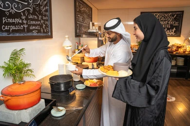 Enjoying time outside the home, spending enjoyable moments on the weekend, a variety of different dishes of delicious and fresh foods and pastries, choosing a breakfast dish, an Emirati Arab couple dressed in traditional Emirati attire serving food from the open buffet in a luxurious restaurant.
