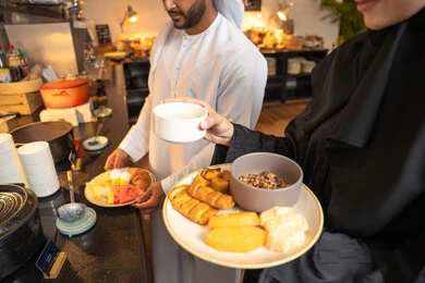 Enjoying time outside the home, spending enjoyable moments on the weekend, a variety of different dishes of delicious and fresh foods and pastries, choosing a breakfast dish, an Emirati Arab couple dressed in traditional Emirati attire serving food from the open buffet in a luxurious restaurant.