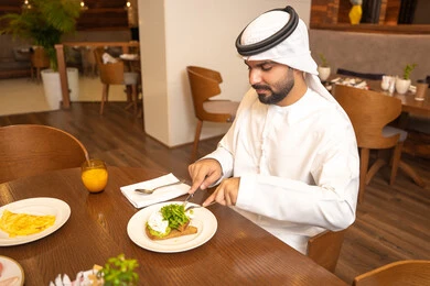 Enjoying outside the home, an Arab Gulf Emirati man wearing a kandura and ghutrah sitting in a luxury restaurant, spending enjoyable times in one of the cafes in the UAE, savoring cold drinks, having breakfast, youth recreational activities.