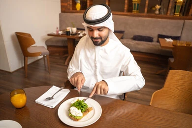 Enjoying outside the home, an Arab Gulf Emirati man wearing a kandura and ghutrah sitting in a luxury restaurant, spending enjoyable times in one of the cafes in the UAE, savoring cold drinks, having breakfast, youth recreational activities.