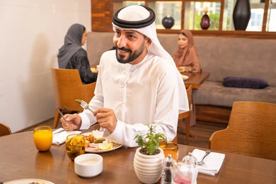 Enjoying outside the home, an Arab Gulf Emirati man wearing a kandura and ghutrah sitting in a luxury restaurant, spending enjoyable times in one of the cafes in the UAE, savoring cold drinks, having breakfast, youth recreational activities.