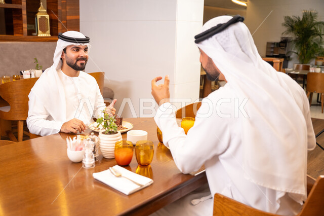 الاستمتاع بتناول الحلا والعصير الطبيعي, تبادل الأحاديث والاخبار, مشاركة الأصدقاء الأوقات الممتعة, بناء علاقات اجتماعية ناجحة, صديقان عربيان خليجيان اماراتيان يرتديان الكندورة والغترة يجلسان في إحدى الكافيهات الفاخرة في المملكة