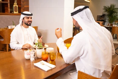 الاستمتاع بتناول الحلا والعصير الطبيعي, تبادل الأحاديث والاخبار, مشاركة الأصدقاء الأوقات الممتعة, بناء علاقات اجتماعية ناجحة, صديقان عربيان خليجيان اماراتيان يرتديان الكندورة والغترة يجلسان في إحدى الكافيهات الفاخرة في المملكة