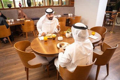 تبادل الأحاديث والاخبار, مشاركة الأصدقاء الأوقات الممتعة, بناء علاقات اجتماعية ناجحة, الاستمتاع بتناول الحلا والعصير الطبيعي, صديقان عربيان خليجيان اماراتيان يرتديان الكندورة والغترة يجلسان في إحدى الكافيهات الفاخرة في المملكة