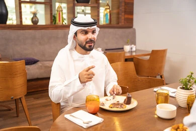 Enjoying outside the home, an Arab Gulf Emirati man wearing a kandura and ghutrah sitting in a luxury restaurant, spending enjoyable times in one of the cafes in the UAE, savoring cold drinks, having breakfast, youth recreational activities.
