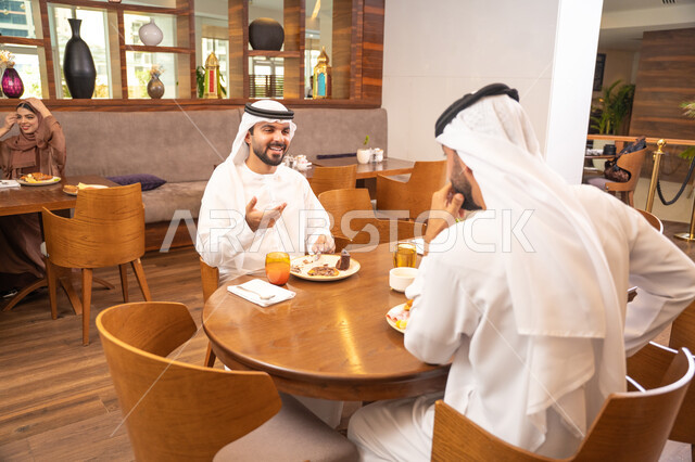 تبادل الأحاديث والاخبار, مشاركة الأصدقاء الأوقات الممتعة, بناء علاقات اجتماعية ناجحة, الاستمتاع بتناول الحلا والعصير الطبيعي, صديقان عربيان خليجيان اماراتيان يرتديان الكندورة والغترة يجلسان في إحدى الكافيهات الفاخرة في المملكة