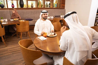 مشاركة الأصدقاء الأوقات الممتعة, بناء علاقات اجتماعية ناجحة, تبادل الأحاديث والاخبار, الاستمتاع بتناول الحلا والعصير الطبيعي, صديقان عربيان خليجيان اماراتيان يرتديان الكندورة والغترة يجلسان في إحدى الكافيهات الفاخرة في المملكة