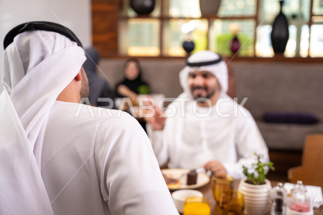تبادل الأحاديث والاخبار, مشاركة الأصدقاء الأوقات الممتعة, بناء علاقات اجتماعية ناجحة, الاستمتاع بتناول الحلا والعصير الطبيعي, صديقان عربيان خليجيان اماراتيان يرتديان الكندورة والغترة يجلسان في إحدى الكافيهات الفاخرة في المملكة