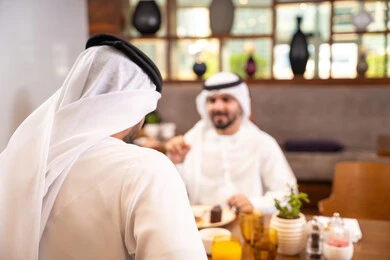 مشاركة الأصدقاء الأوقات الممتعة, بناء علاقات اجتماعية ناجحة, تبادل الأحاديث والاخبار, الاستمتاع بتناول الحلا والعصير الطبيعي, صديقان عربيان خليجيان اماراتيان يرتديان الكندورة والغترة يجلسان في إحدى الكافيهات الفاخرة في المملكة