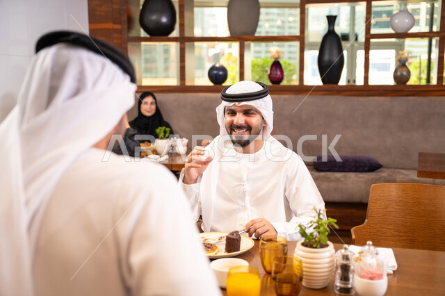 مشاركة الأصدقاء الأوقات الممتعة, بناء علاقات اجتماعية ناجحة, تبادل الأحاديث والاخبار, الاستمتاع بتناول الحلا والعصير الطبيعي, صديقان عربيان خليجيان اماراتيان يرتديان الكندورة والغترة يجلسان في إحدى الكافيهات الفاخرة في المملكة