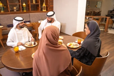 A fun and social gathering, discussions and family dialogues at the dining table, an Emirati Arab Gulf family dressed in traditional attire sitting in a luxurious restaurant, enjoying delicious meals, spending enjoyable family time on the weekend, friendships, and building social relationships.