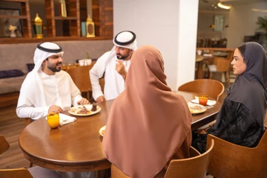 Spending enjoyable family time on the weekend, having delicious meals, a fun gathering and getting to know each other, discussions and family dialogues at the dining table, an Emirati Arab Gulf family dressed in traditional attire sitting in a luxurious restaurant, friendships and building social relationships.