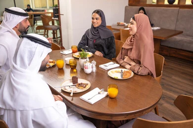 A fun and social gathering, discussions and family dialogues at the dining table, an Emirati Arab Gulf family dressed in traditional attire sitting in a luxurious restaurant, enjoying delicious meals, spending enjoyable family time on the weekend, friendships, and building social relationships.
