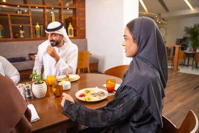 Spending enjoyable family time on the weekend, having delicious meals, a fun gathering and getting to know each other, discussions and family dialogues at the dining table, an Emirati Arab Gulf family dressed in traditional attire sitting in a luxurious restaurant, friendships and building social relationships.