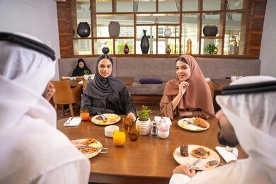 A fun and social gathering, discussions and family dialogues at the dining table, an Emirati Arab Gulf family dressed in traditional attire sitting in a luxurious restaurant, enjoying delicious meals, spending enjoyable family time on the weekend, friendships, and building social relationships.