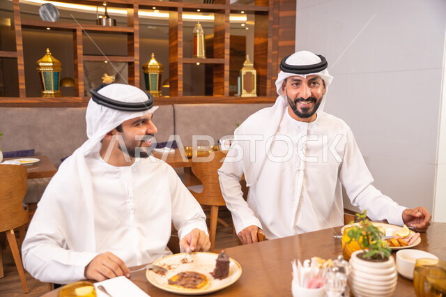 Enjoying desserts and natural juice, exchanging conversations and news, sharing enjoyable moments with friends, building successful social relationships, two Emirati Gulf Arab friends wearing kandura and ghutrah sitting in one of the luxurious cafes in the kingdom.