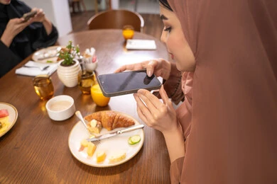 Spending a good time at the restaurant, using a modern and advanced technical device, a close-up image of a veiled Emirati Gulf Arab woman wearing an abaya, a young Saudi woman taking a souvenir photo of the food dishes with her mobile phone, enjoying hot drinks and desserts in restaurants and cafes in the UAE.