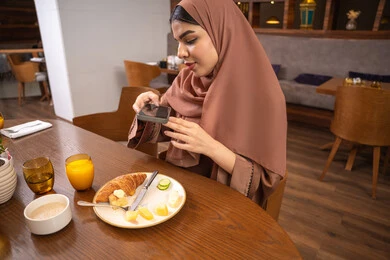 Spending a good time at the restaurant, using a modern and advanced technical device, a close-up image of a veiled Emirati Gulf Arab woman wearing an abaya, a young Saudi woman taking a souvenir photo of the food dishes with her mobile phone, enjoying hot drinks and desserts in restaurants and cafes in the UAE.