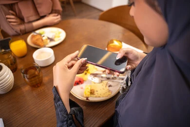 Using a modern and advanced technical device, having a good time at the restaurant, a close-up image of a veiled Emirati Gulf Arab woman wearing an abaya, a young Saudi woman taking a commemorative photo of the food dishes with her mobile phone, enjoying delicious food in restaurants and cafes in the UAE.
