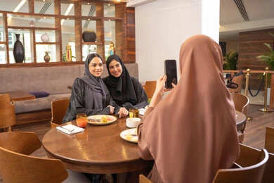 Taking commemorative photos using a mobile phone, a fun and social gathering, building social relationships, a fine restaurant in the UAE, delicious foods and meals, having an enjoyable conversation among friends, Arab Gulf Emirati friends sitting in one of the luxurious restaurants, a meeting on the weekend.