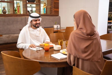 Gestures of happiness and joy, the concept of a happy family, love and affection between spouses, spending enjoyable time outside the home on the weekend, a fine restaurant in the United Arab Emirates, an Arab Gulf Emirati husband wearing the kandura and ghutrah sitting with his wife in the restaurant, enjoying delicious food.