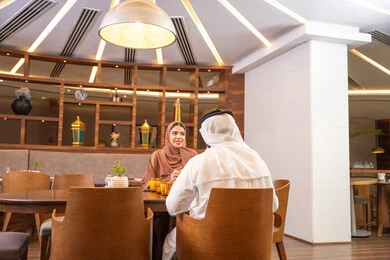 The concept of a happy family, love and affection between spouses, spending enjoyable time outside the home on the weekend, a fine restaurant in the United Arab Emirates, an Arab Gulf Emirati husband wearing the kandura and ghutrah sitting with his wife in the restaurant, enjoying delicious food.