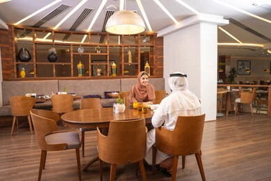 A fine restaurant in the United Arab Emirates, gestures of happiness and joy, the concept of a happy family, love and affection between spouses, spending enjoyable time outside the home on the weekend, an Emirati Gulf Arab husband wearing the kandura and ghutrah sitting with his wife in the restaurant, enjoying delicious dishes.