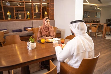 The concept of a happy family, love and affection between spouses, spending enjoyable time outside the home on the weekend, a fine restaurant in the United Arab Emirates, an Arab Gulf Emirati husband wearing the kandura and ghutrah sitting with his wife in the restaurant, enjoying delicious food.