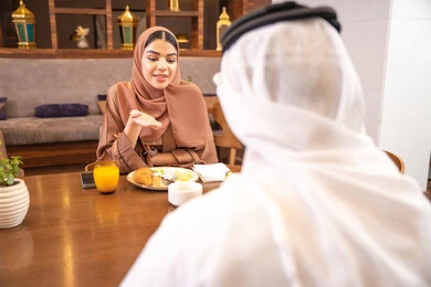 مطعم راقي في الامارات العربية المتحدة، ايماءات السعادة والسرور، مفهوم الاسره السعيده, المحبة والمودة بين الزوجين، قضاء وقت ممتعه خارج المنزل في عطلة نهاية الاسبوع, زوج عربي خليجي اماراتي يرتدي الكندورة والغترة يجلس مع زوجته في المطعم, تناول المأكولات الشهية