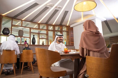 Gestures of happiness and joy, the concept of a happy family, love and affection between spouses, spending enjoyable time outside the home on the weekend, a fine restaurant in the United Arab Emirates, an Arab Gulf Emirati husband wearing the kandura and ghutrah sitting with his wife in the restaurant, enjoying delicious food.