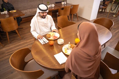 مفهوم الاسره السعيده, المحبة والمودة بين الزوجين، قضاء وقت ممتعه خارج المنزل في عطلة نهاية الاسبوع, مطعم راقي في الامارات العربية المتحدة، زوج عربي خليجي اماراتي يرتدي الكندورة والغترة يجلس مع زوجته في المطعم, تناول المأكولات الشهية