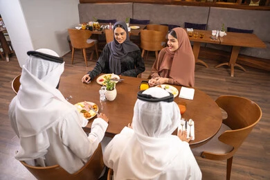 Spending enjoyable family time on the weekend, a fun gathering and getting to know each other, discussions and family dialogues at the dining table, an Emirati Arab Gulf family dressed in traditional attire sitting in a luxurious restaurant, enjoying delicious meals, friendships, and building social relationships.