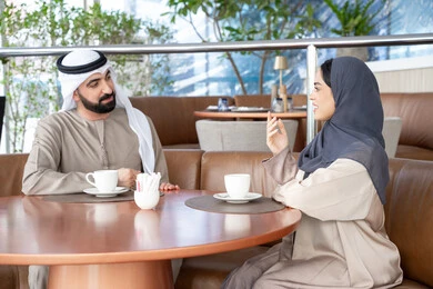 التفاهم والترابط الفكري بين الزوجين، شرب القهوة العربية، قضاء أوقات عائلية ممتعة، رجل عربي خليجي اماراتي يرتدي الكندورة والغترة يجلس فى احد المقاهي يقوم بتبادل الأحاديث مع زوجته في الهواء الطلق