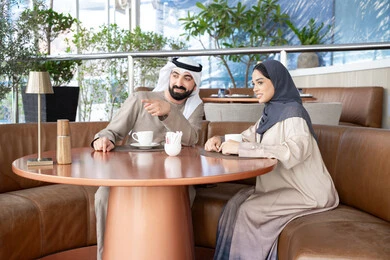 إيماءة تدل على شيء محدد، مشاعر المودة والترابط بين الزوجين، قضاء أوقات ممتعة، رجل عربي خليجي اماراتي يرتدي الكندورة و الغتره يجلس فى احد المقاهي برفقة زوجته ، تعابير السعادة و السرور