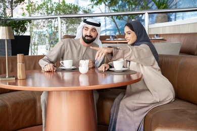 توجيه النظر إلى شيء محدد، مشاعر المودة والترابط بين الزوجين، قضاء أوقات ممتعة، رجل عربي خليجي اماراتي يرتدي الكندورة و الغتره يجلس فى احد المقاهي برفقة زوجته ، ايماءات السعادة و السرور