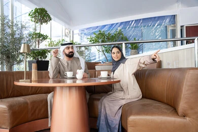 لفت الانتباه نحو شيء معين، مفهوم المودة والالفة ، قضاء أوقات ممتعة، رجل عربي خليجي اماراتي يرتدي الكندورة و الغتره يجلس فى احد المقاهي برفقة زوجته ، ايماءات السعادة و السرور