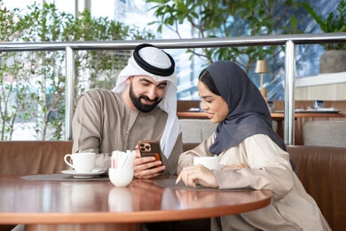 Browsing news and content on social media platforms, having a good time outdoors, entertainment and fun during the holiday, the concept of affection and intimacy, an Arab Gulf Emirati man wearing a kandura and ghutrah sitting in a café with his wife watching a mobile phone, gestures of happiness and joy.