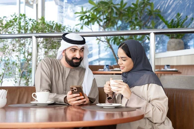 Following news on social media, spending enjoyable time outdoors, entertainment and fun during the holiday, the concept of affection and intimacy, enjoying favorite drinks, an Arab Gulf Emirati man wearing a kandura and ghutrah sitting in a café with his wife watching a mobile phone, gestures of happiness and joy.