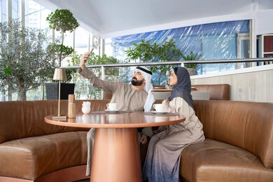 التواصل مع الاهل والاصدقاء، التقاط صور التذكارية عبر الجوال، الأجواء العائلية السعيدة، توثيق اللحظات السعيدة ، تقوية العلاقات الزوجية وروابط الحب والمودة، زوجان عربيان خليجيان اماراتيان يرتديان الزي التقليدي يجلسان فى احد المقاهي بالامارات العربية
