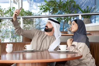 Integrating modern technologies and techniques into daily life, capturing memorable photos via mobile, happy family atmospheres, documenting joyful moments, strengthening marital relationships and bonds of love and affection, an Emirati Gulf Arab couple dressed in traditional attire sitting in one of the cafes in the United Arab Emirates.