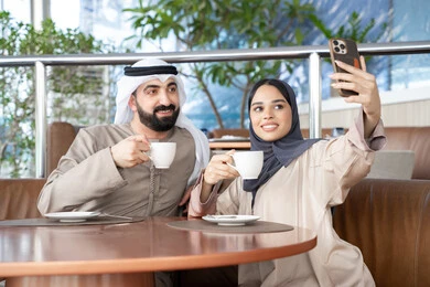 توثيق اللحظات السعيدة ، تقوية العلاقات الزوجية وروابط الحب والمودة، التقاط صور السيلفي التذكارية باستخدام الهاتف المحمول، زوجان عربيان خليجيان اماراتيان يرتديان الزي التقليدي يجلسان فى احد المقاهي بالامارات العربية، تناول المشروبات المفضلة
