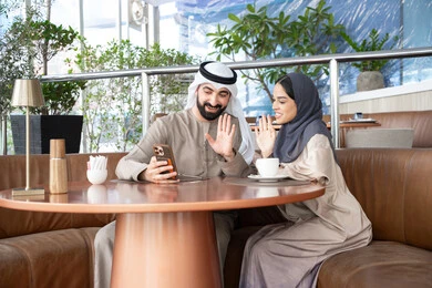 Spending time talking with friends and family, a welcoming gesture by raising the hand, making video calls online, using modern technological devices, an Emirati Gulf Arab man wearing a kandura and ghutrah sitting in a café with his wife using a mobile phone, expressions of happiness and joy.