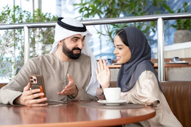 تحية ودية برفع كف اليد ، التحدث والتواصل مع العائلة والأصدقاء، إجراء مكالمات الفيديو عبر الانترنت، استخدام اجهزة تقنية حديثة، رجل عربي خليجي اماراتي يرتدي الكندورة و الغتره يجلس فى احد المقاهي برفقة زوجته يستخدمان الهاتف المحمول ، ايماءات السعادة و السرور