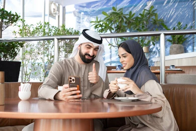 تعابير الموافقة و القبول، التواصل الاجتماعي مع الأصدقاء والعائلة، إجراء مكالمات الفيديو عبر الانترنت، الاستمتاع بتناول المشروب المفضل، رجل عربي خليجي اماراتي يرتدي الكندورة و الغتره يجلس فى احد المقاهي برفقة زوجته يستخدمان الهاتف المحمول ، ايماءات السعادة و السرور