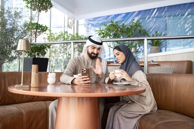 Raising the hand to greet and welcome, socializing with friends and family, making video calls online, enjoying a favorite drink, an Emirati Gulf Arab man wearing a kandura and ghutrah sitting in a café with his wife using a mobile phone, gestures of happiness and joy.