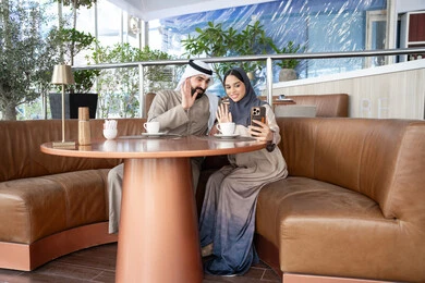 تبادل الأحاديث مع الأقارب والأصدقاء، رفع كف الايد بتعابير السلام و التحية ، إجراء مكالمات الفيديو عبر الانترنت، استخدام اجهزة تقنية حديثة، رجل عربي خليجي اماراتي يرتدي الكندورة و الغتره يجلس فى احد المقاهي برفقة زوجته يستخدمان الهاتف المحمول ، ايماءات السعادة و السرور