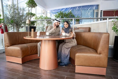 رفع كف الايد بتعابير السلام و التحية ، التحدث والتواصل مع العائلة والأصدقاء، إجراء مكالمات الفيديو عبر الانترنت، استخدام اجهزة تقنية حديثة، رجل عربي خليجي اماراتي يرتدي الكندورة و الغتره يجلس فى احد المقاهي برفقة زوجته يستخدمان الهاتف المحمول ، ايماءات السعادة و السرور